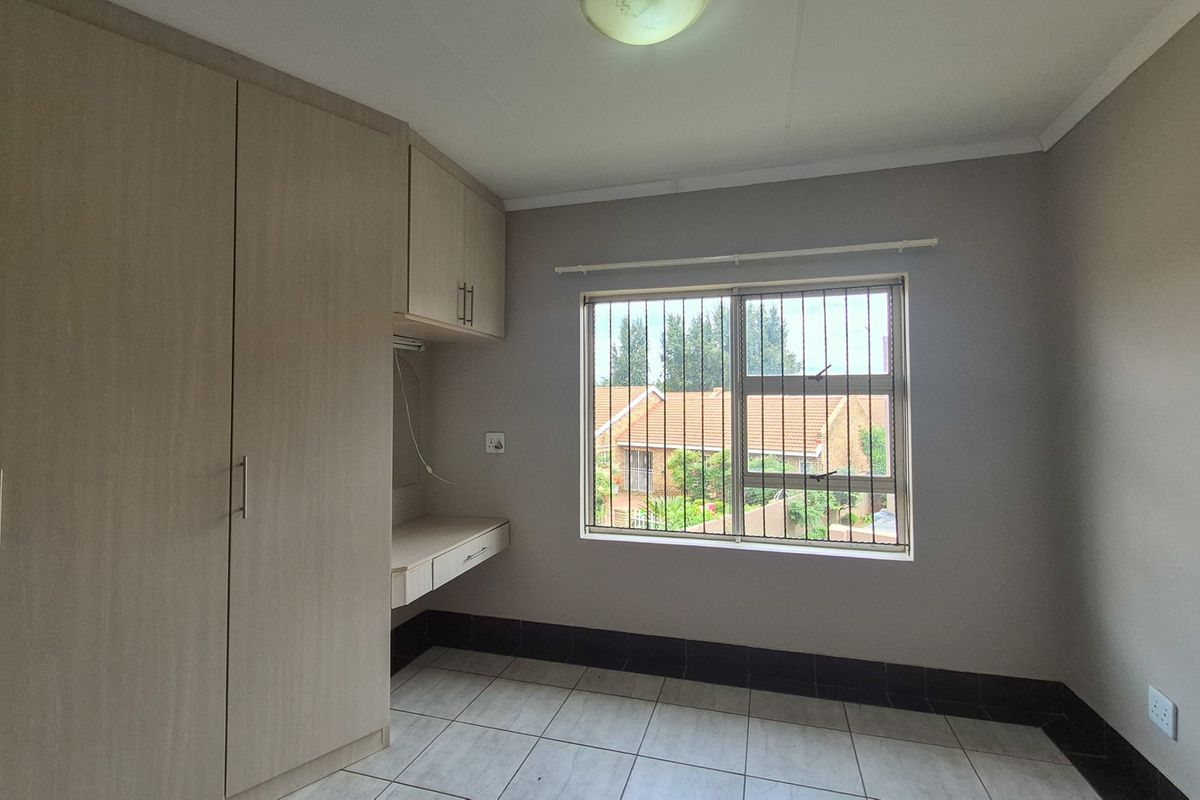 A clean, empty bedroom featuring neutral tiled flooring, built-in cupboards, a integrated desk area, and a window fitted with security bars.