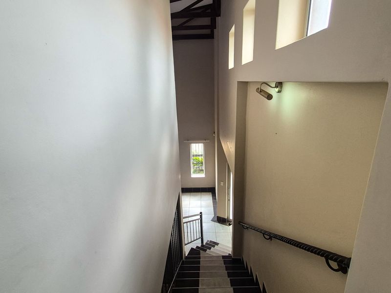A high-ceilinged hallway featuring a staircase with a wrought iron handrail, exposed timber beams, and tiled flooring.
