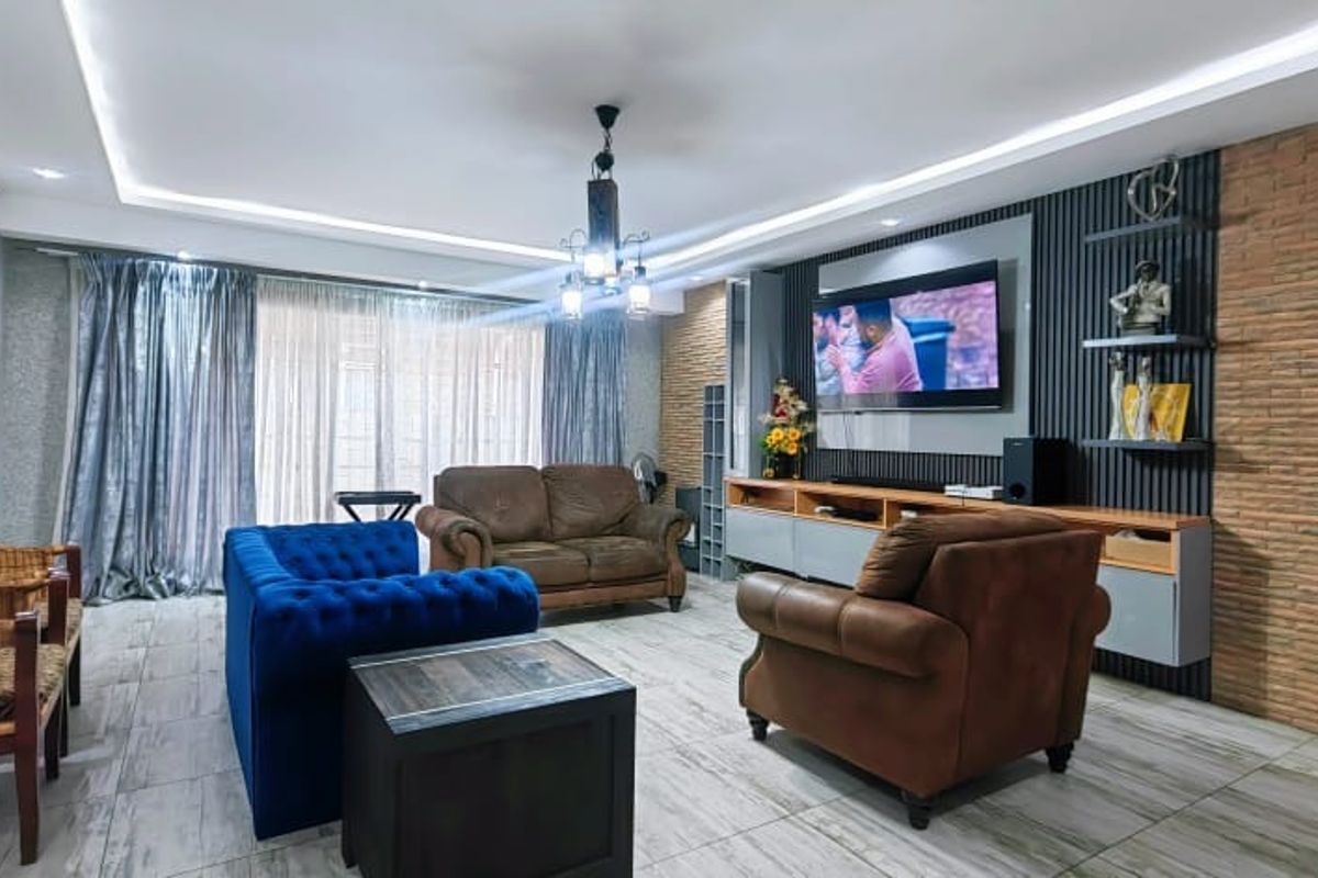 A well-maintained living room featuring a modern media wall with vertical slats, exposed brick accents, recessed cove lighting, and contemporary tiled flooring.