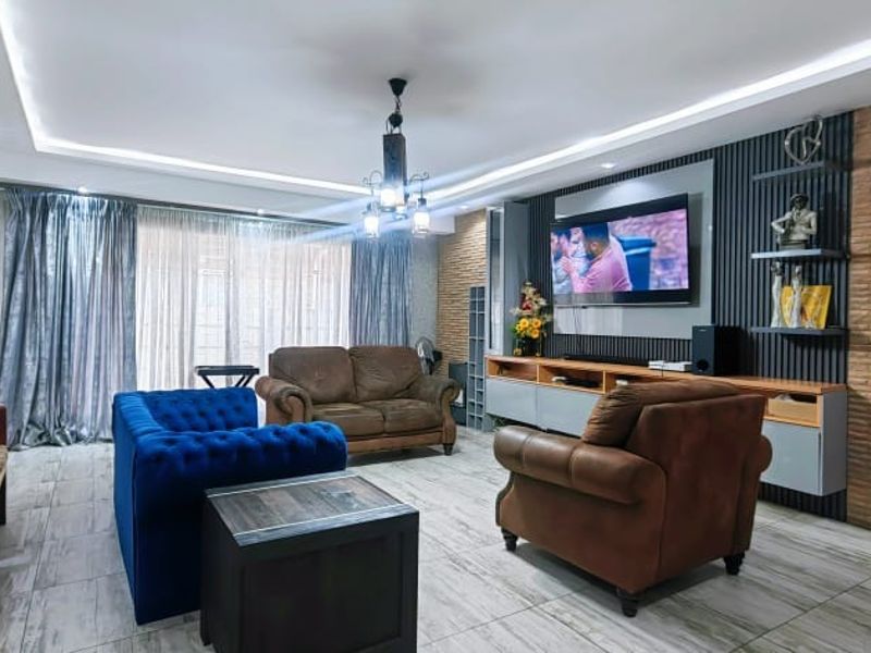 A well-maintained living room featuring a modern media wall with vertical slats, exposed brick accents, recessed cove lighting, and contemporary tiled flooring.