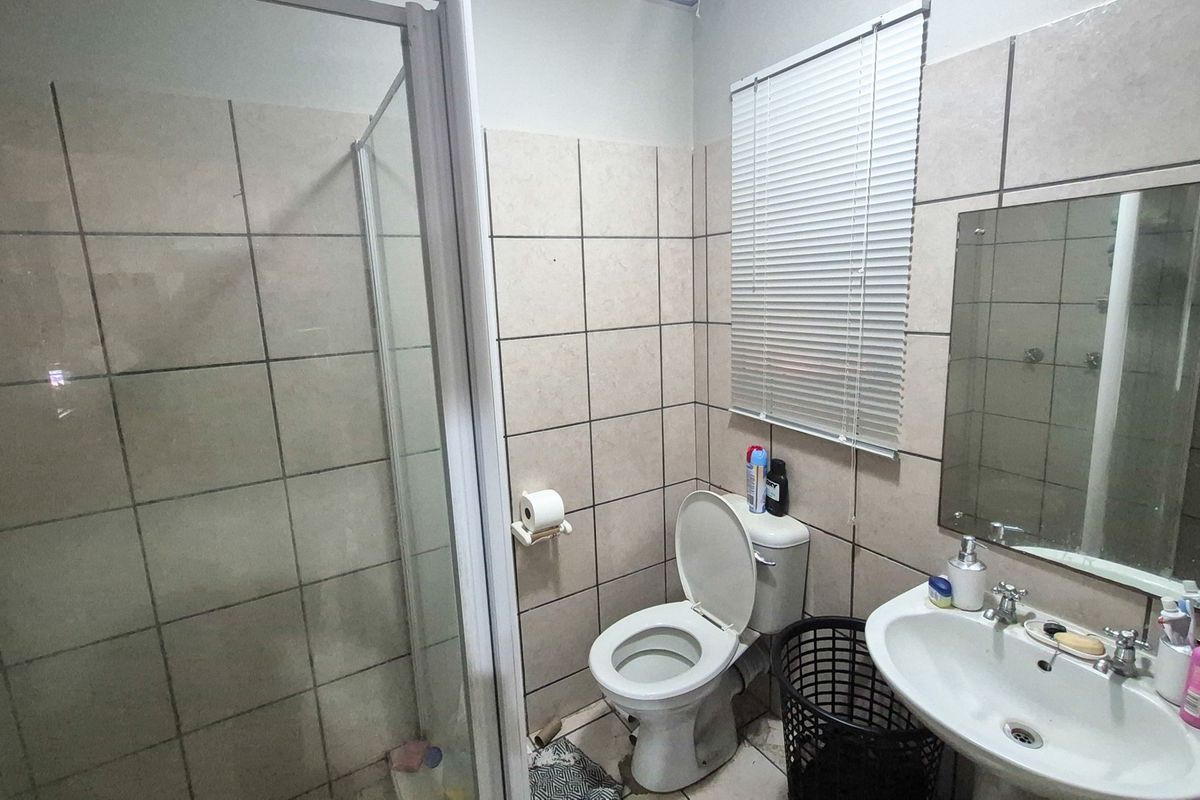 A functional bathroom featuring a shower enclosure, pedestal basin, and toilet, finished with neutral ceramic wall tiles and a window blind.