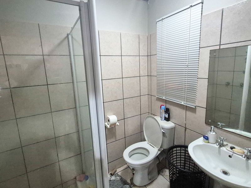 A functional bathroom featuring a shower enclosure, pedestal basin, and toilet, finished with neutral ceramic wall tiles and a window blind.