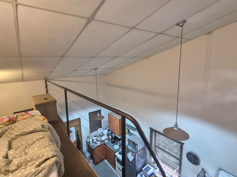 A mezzanine bedroom level overlooking the kitchen area, featuring a metal safety balustrade and suspended pendant lighting under a drop ceiling.