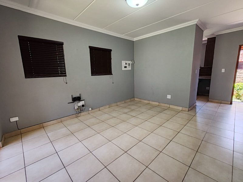 An empty, open-plan living area featuring neutral grey walls, tiled flooring, and venetian blinds, with a clear view leading toward the kitchen area.