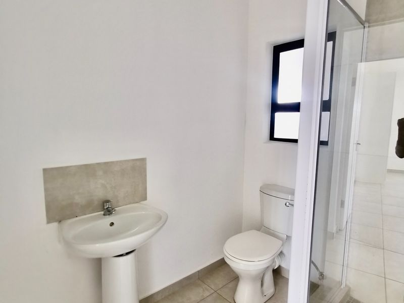 A clean, modern bathroom featuring a pedestal basin, a standard toilet, and a glass-enclosed shower with neutral floor tiling.