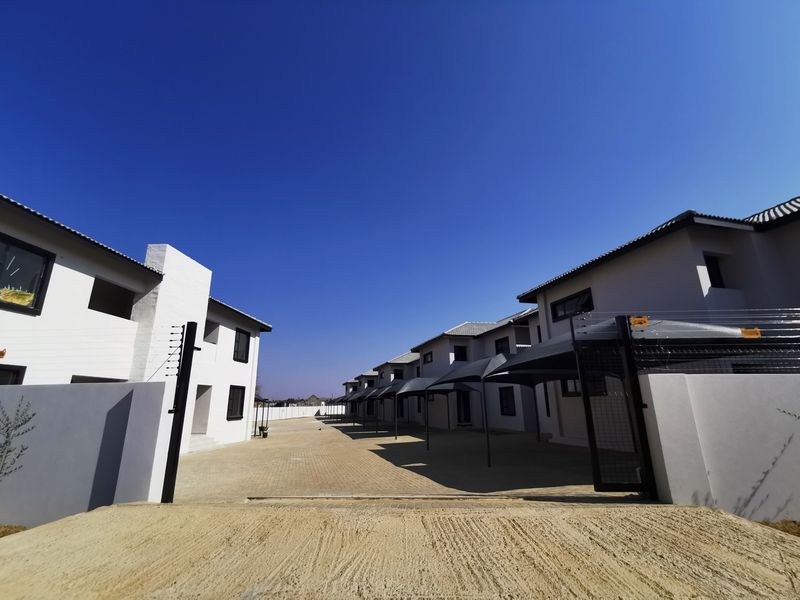 A wide-angle view of an apartment complex exterior featuring a paved driveway, covered parking bays, and secure perimeter walling with electric fencing.