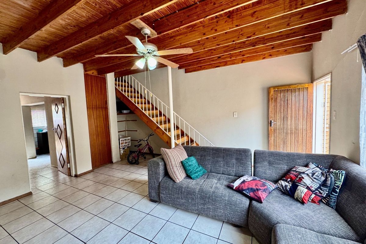 Living room featuring exposed timber beam ceilings, tiled flooring, and a staircase with metal railing, offering a functional and dated layout.