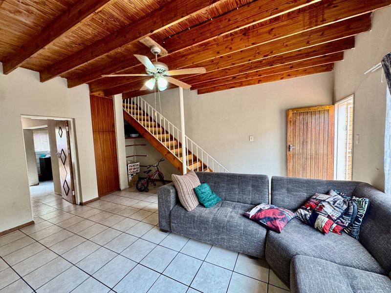 Living room featuring exposed timber beam ceilings, tiled flooring, and a staircase with metal railing, offering a functional and dated layout.