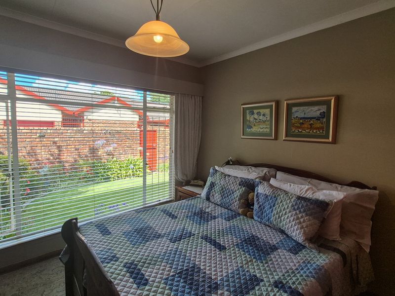 A well-lit bedroom featuring large windows with venetian blinds, neutral wall tones, and carpeted flooring, offering a comfortable and functional living space.