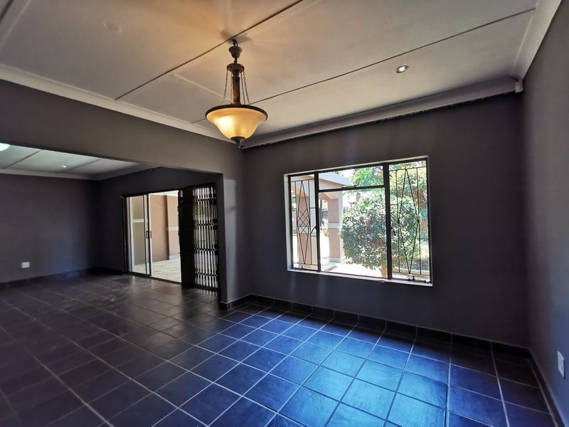 An empty, spacious living area featuring dark tiled flooring, a classic pendant light, and large windows with security bars, leading to an outdoor patio.