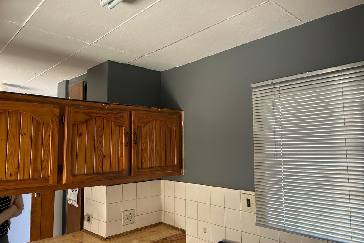A kitchen area featuring traditional wooden cabinetry, a tiled backsplash, and venetian blinds, showing signs of wear and dated finishes.