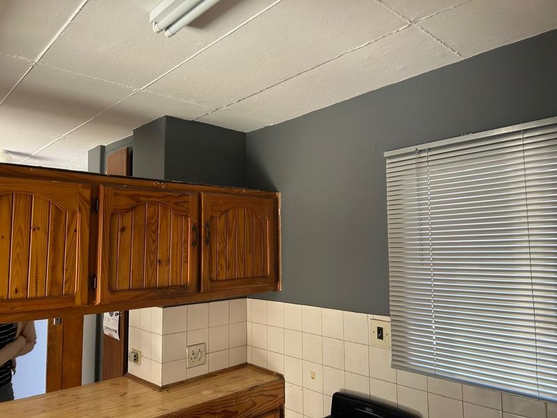 A kitchen area featuring traditional wooden cabinetry, a tiled backsplash, and venetian blinds, showing signs of wear and dated finishes.