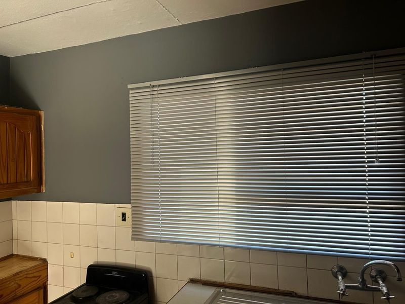 A kitchen area featuring a tiled backsplash, built-in wooden cabinetry, a stainless steel sink, and an electric stove, finished with venetian blinds.