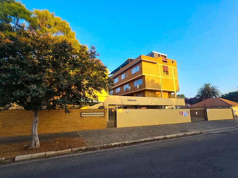 Front exterior view of the Sherada apartment complex, featuring a brick boundary wall, secure gated entrance, and paved driveway access.