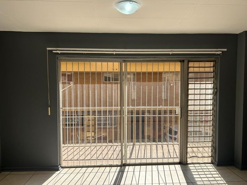 An empty living room featuring tiled flooring and a large sliding glass door with an integrated security gate, providing access to the exterior.