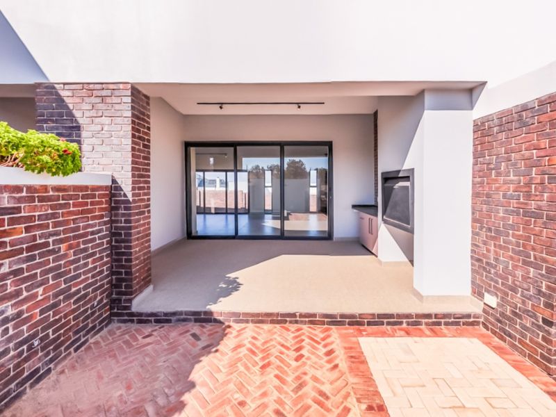 A covered patio area featuring brick paving, a built-in braai, and sliding glass doors leading into the interior living space.
