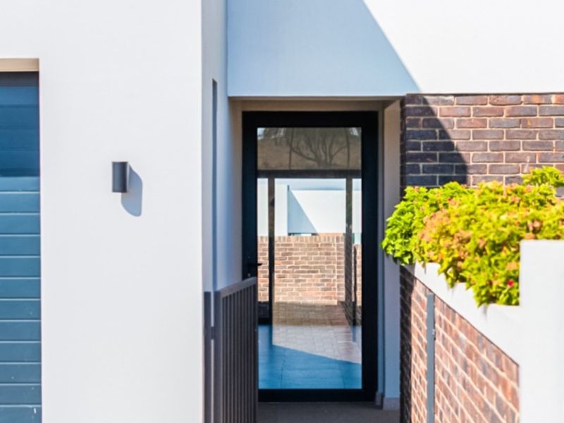 Modern front entrance featuring a clean white facade, brick accents, a secure metal gate, and a glass door leading into the property.