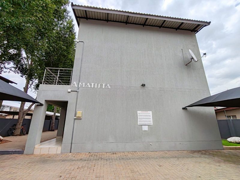 Exterior view of the Amatista apartment complex building featuring a paved driveway, metal balcony railing, and mounted satellite dish.