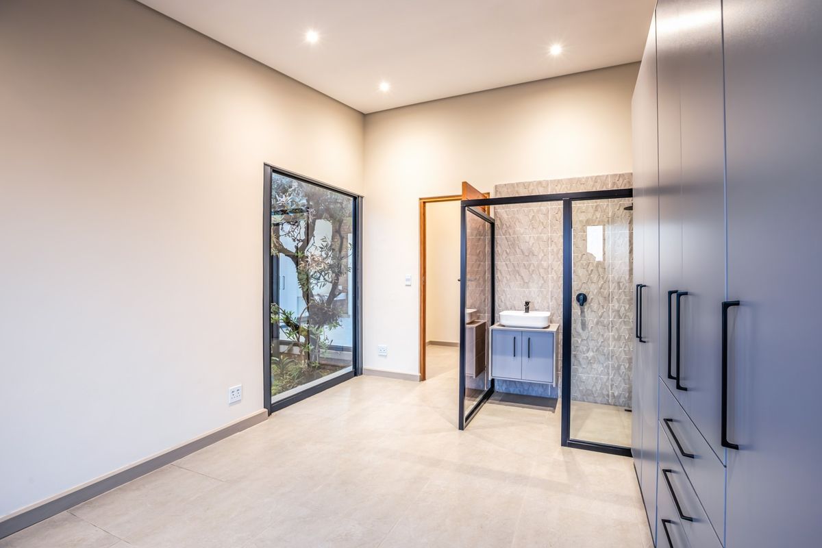 Modern bedroom featuring built-in cupboards, tiled flooring, recessed lighting, and an open-plan en-suite shower with a vanity basin and large feature window.