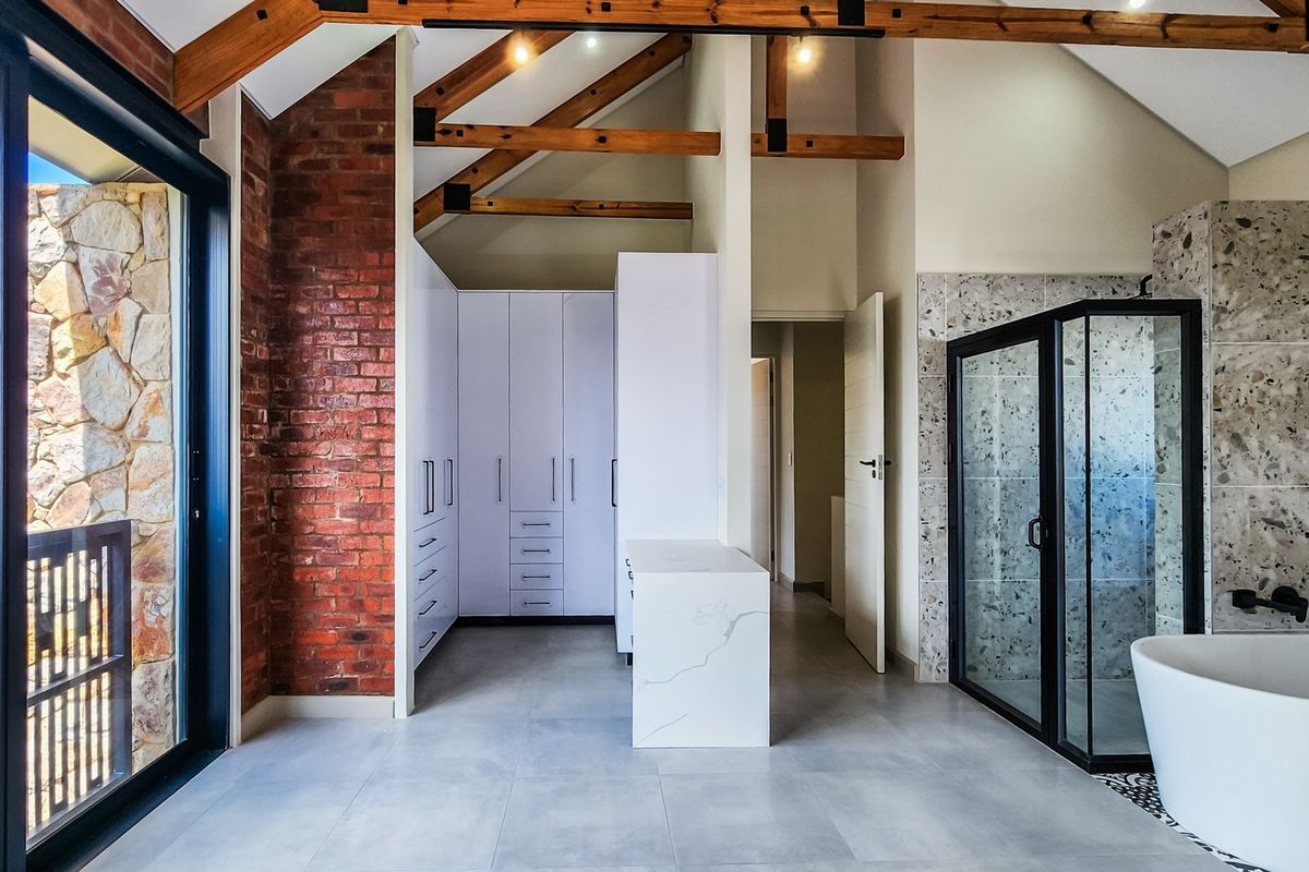 Modern luxury bathroom featuring exposed timber trusses, brick feature wall, freestanding tub, walk-in shower, and custom cabinetry with a stone-topped island.