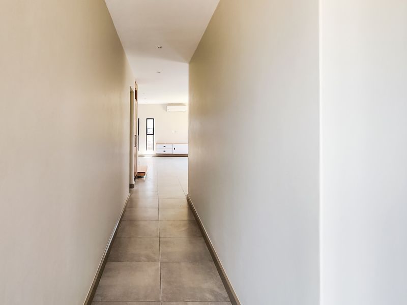 A clean, neutral hallway featuring large format floor tiles and recessed lighting, leading into an open-plan living area with air conditioning.
