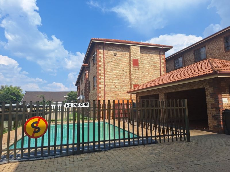 Exterior view of an apartment complex featuring a swimming pool, brick building facade, and secure metal fencing.
