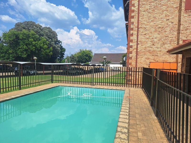 A communal swimming pool area featuring brick paving and metal perimeter fencing, situated adjacent to a face brick apartment building and covered parking.
