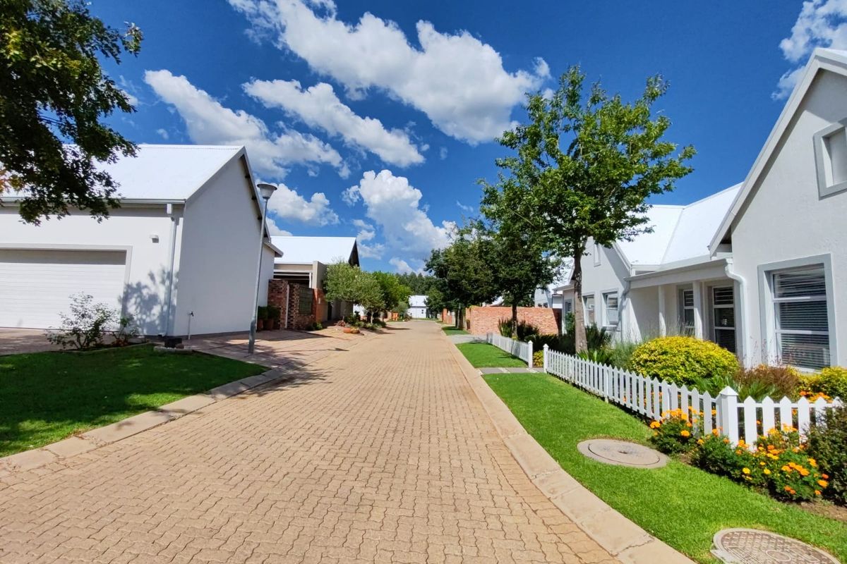 A well-maintained residential street scene featuring paved driveways, white picket fencing, and manicured garden verges within a clean, modern estate setting.