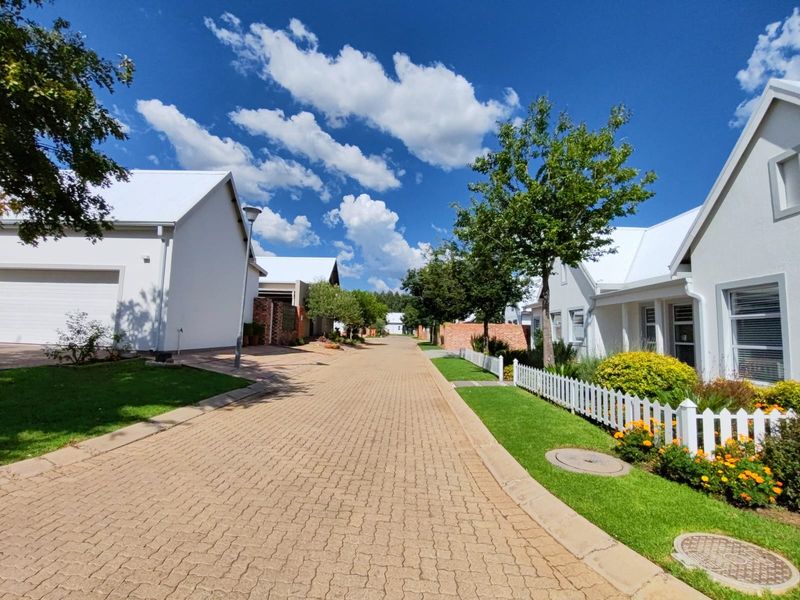 A well-maintained residential street scene featuring paved driveways, white picket fencing, and manicured garden verges within a clean, modern estate setting.