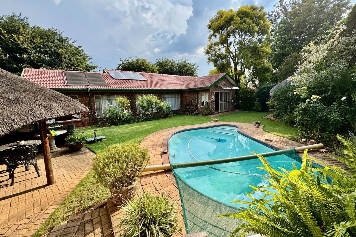 Rear garden featuring a swimming pool, brick-paved patio area, and a thatched lapa, set against a brick house with solar panels on the roof.