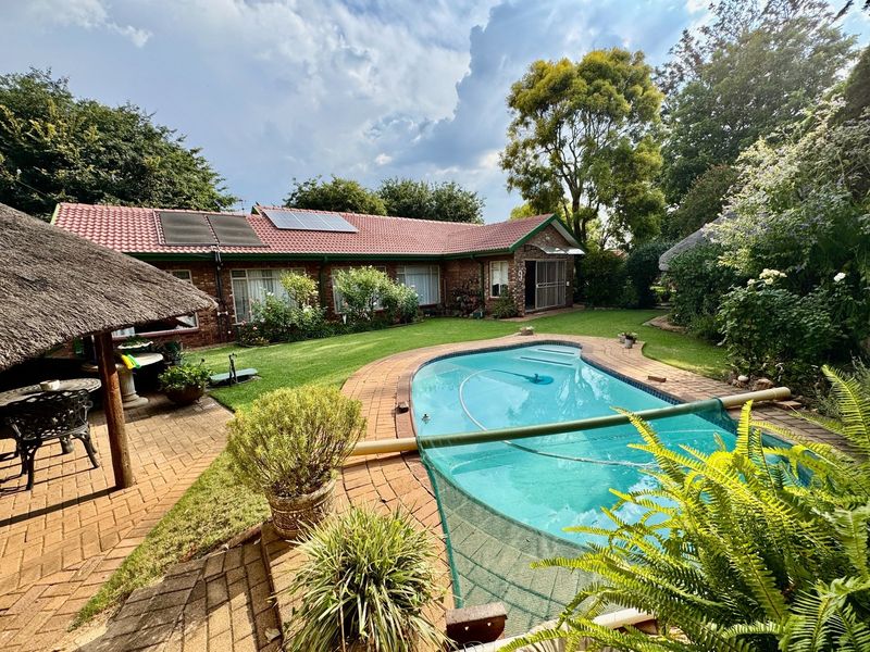 Rear garden featuring a swimming pool, brick-paved patio area, and a thatched lapa, set against a brick house with solar panels on the roof.