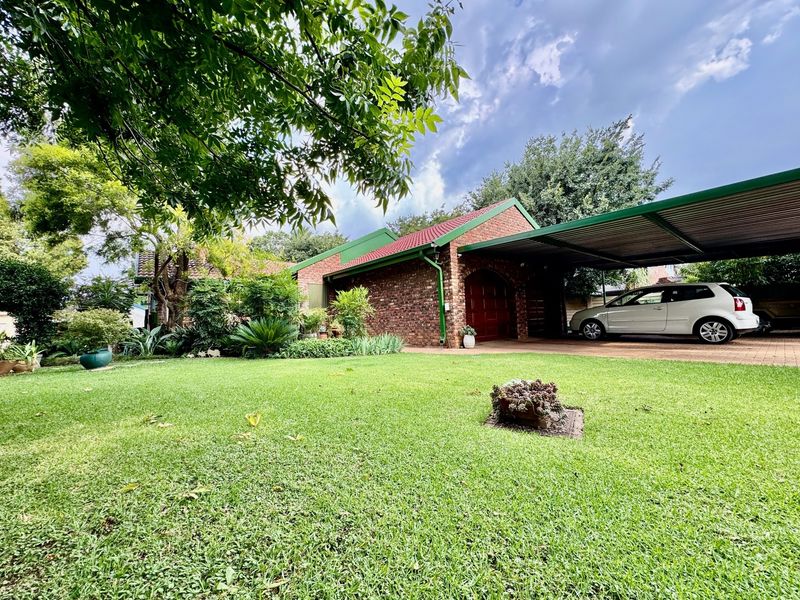 A well-maintained brick house featuring a spacious front garden, a paved driveway, and a covered carport area.