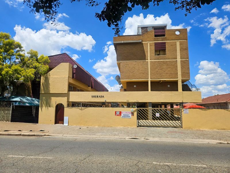 Street-facing view of the Sherada apartment complex, featuring a brick facade, secure gated entrance, and perimeter walling with security fencing.