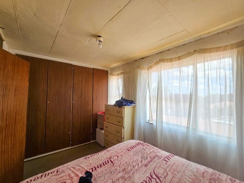 A bedroom featuring built-in wooden cupboards, a large window allowing natural light, and a tiled ceiling. The room requires cosmetic updates to finishes.