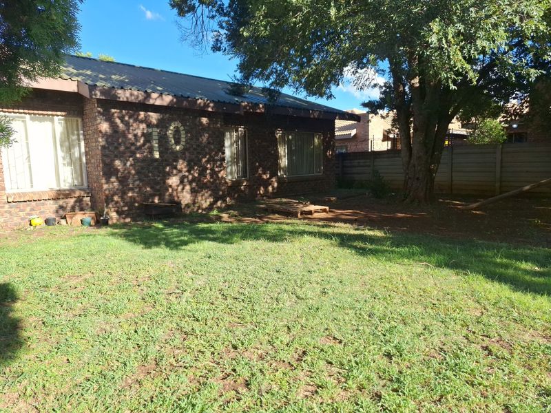 A rear view of a face brick house featuring a metal roof, set within a garden with a large mature tree and boundary wall.