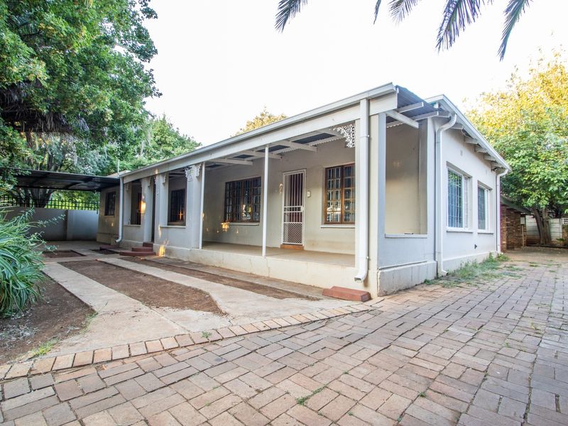 Front elevation of a single-story house featuring a covered veranda, paved driveway, and established garden surroundings.