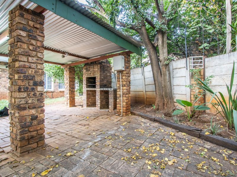 Covered outdoor patio area featuring a built-in brick braai, paved flooring, and brick support pillars, set within a garden environment.