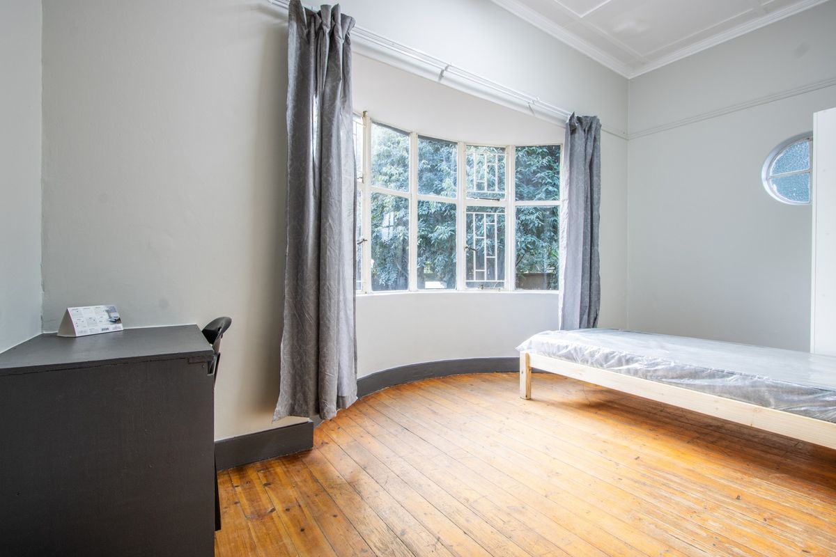 A bright bedroom featuring original hardwood flooring, a distinctive bay window, and a unique circular window, offering a clean and neutral canvas.