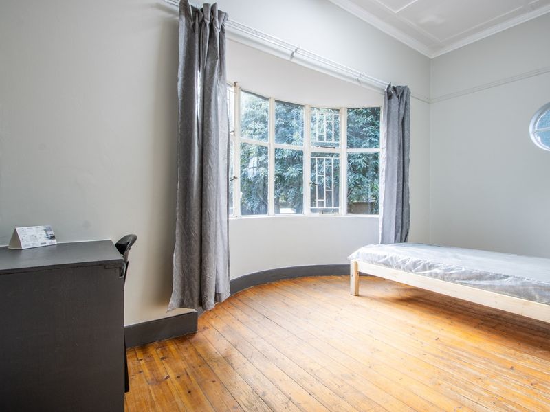 A bright bedroom featuring original hardwood flooring, a distinctive bay window, and a unique circular window, offering a clean and neutral canvas.