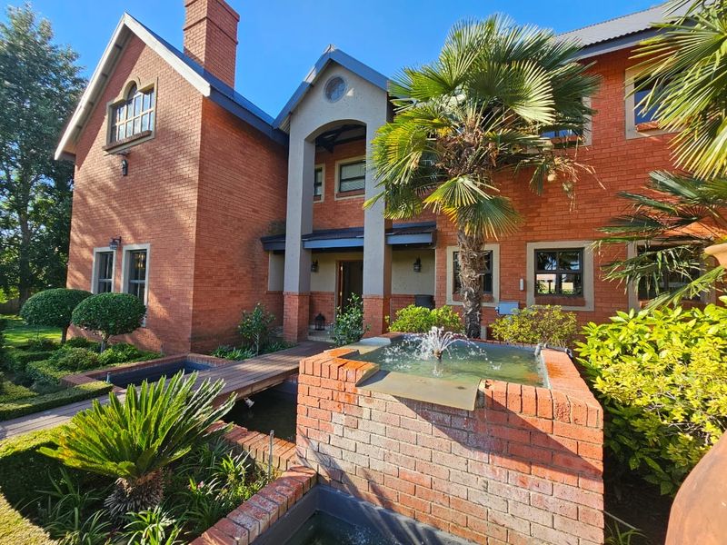 A well-maintained double-story brick residence featuring a decorative water fountain, a wooden footbridge over a pond, and established garden landscaping.