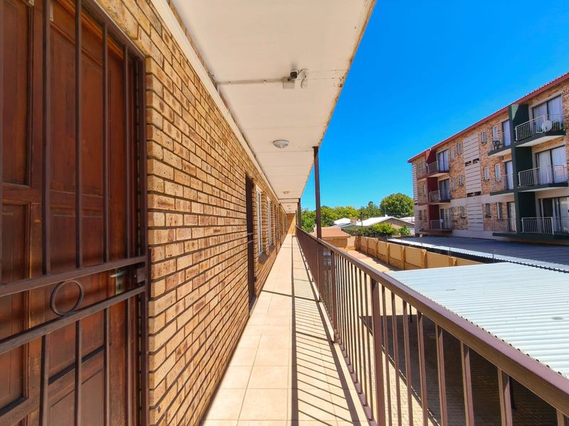 An exterior apartment balcony featuring brick wall finishes, tiled flooring, and a metal safety balustrade overlooking the complex grounds.