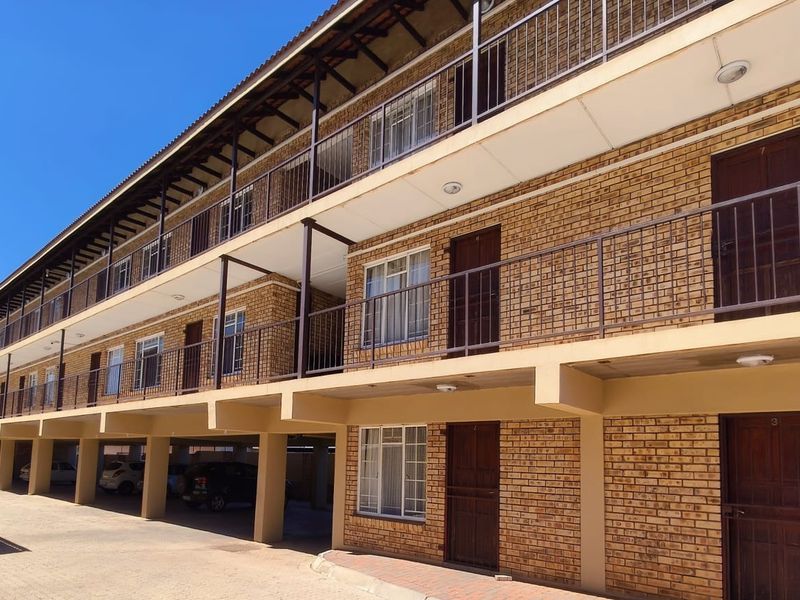 A multi-story apartment building featuring a face brick facade, open-air walkways with metal railings, and ground-level covered parking bays.
