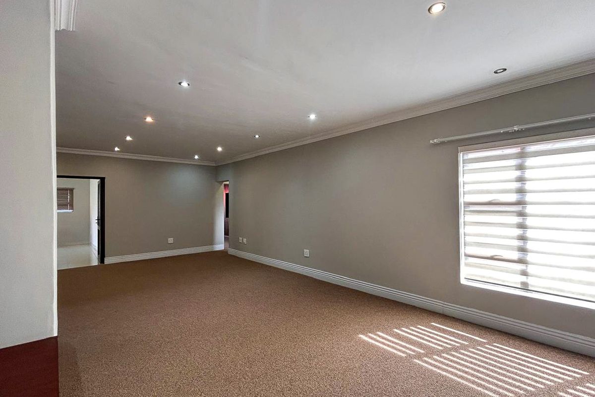 A spacious, empty living room featuring neutral wall tones, carpeted flooring, recessed downlighting, and large windows with fitted roller blinds.