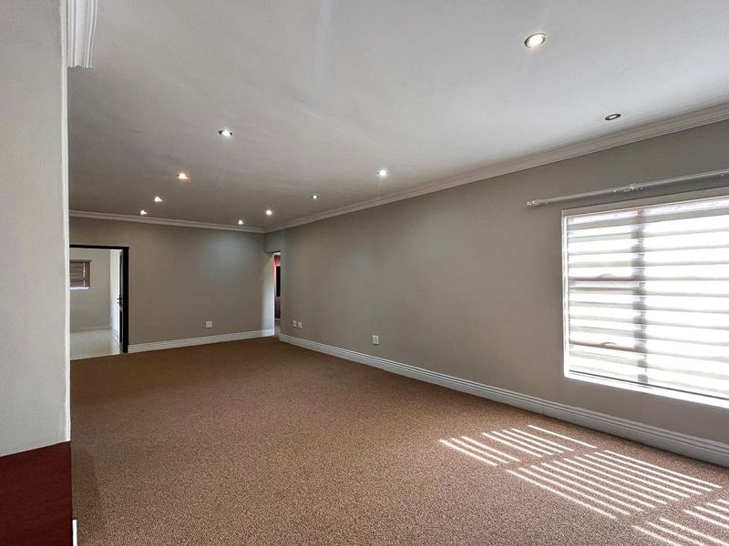 A spacious, empty living room featuring neutral wall tones, carpeted flooring, recessed downlighting, and large windows with fitted roller blinds.