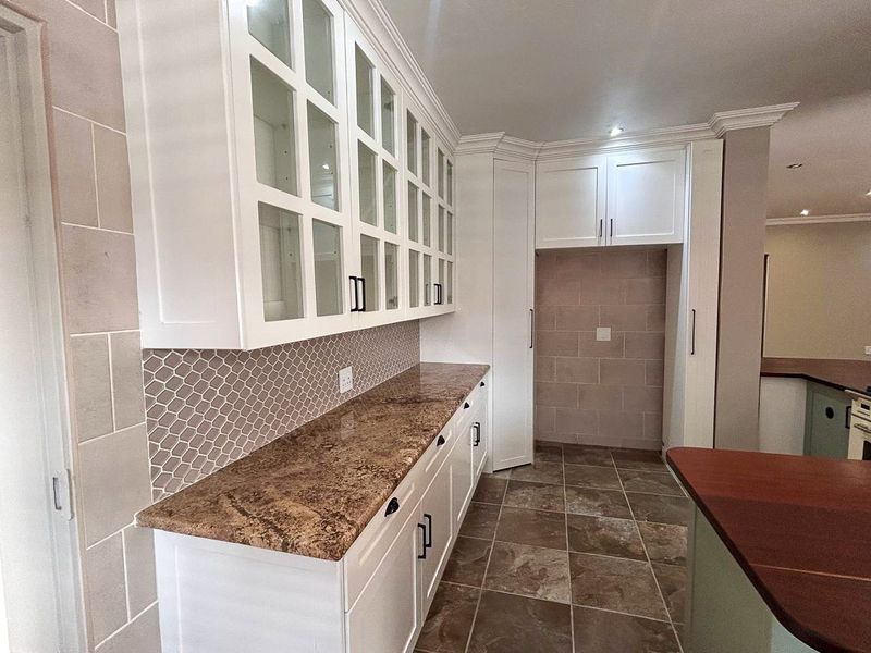 A clean, modern kitchen featuring white cabinetry with glass-fronted uppers, granite countertops, a decorative tiled backsplash, and neutral ceramic floor tiles.