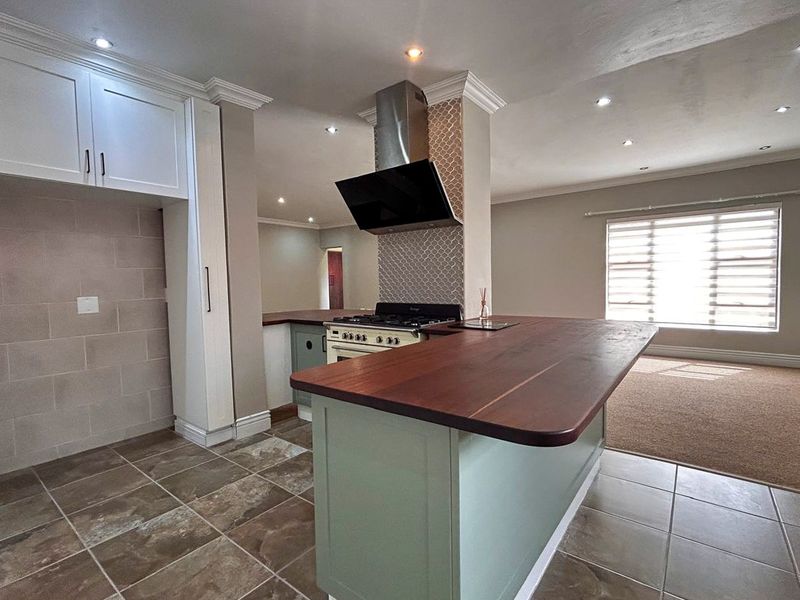 A spacious, modern kitchen featuring a central island with a wooden countertop, gas stove, extractor fan, and neutral cabinetry, seamlessly flowing into the living area.