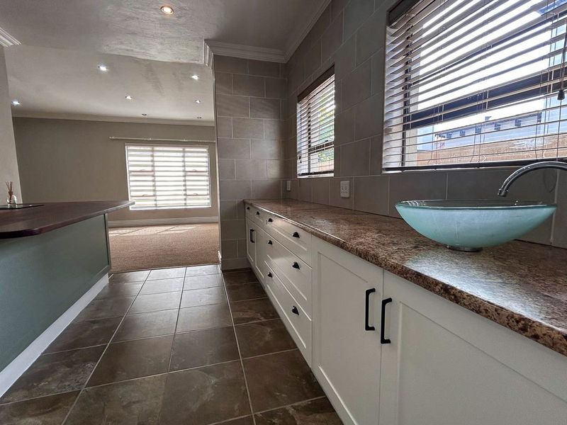 A clean, modern scullery featuring white built-in cabinetry, granite countertops, a vessel sink, and tiled flooring, with an adjacent view into the living area.
