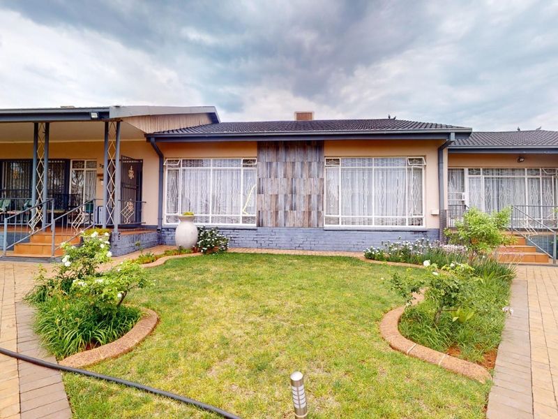 A well-maintained rear exterior view featuring a paved courtyard, landscaped garden beds, and a covered patio area with security gates.