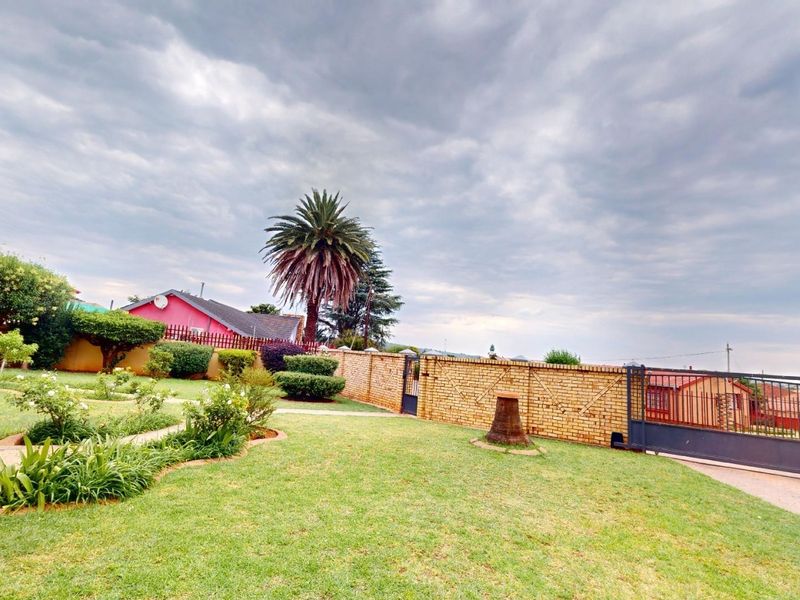 A well-maintained garden area featuring a brick boundary wall, metal sliding gate, and paved pathways set within a landscaped lawn.