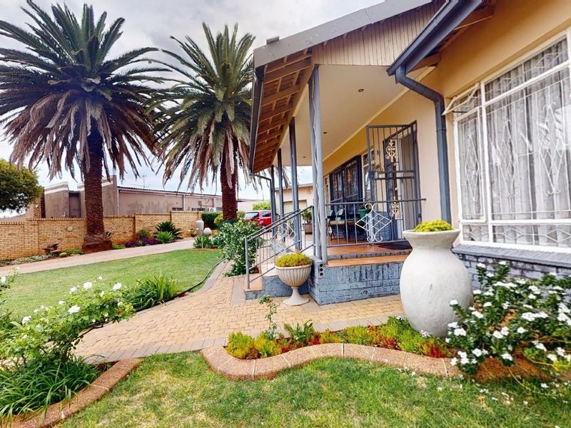 A well-maintained front exterior featuring a covered porch, brick paved driveway, and established garden with mature palm trees and secure boundary walling.